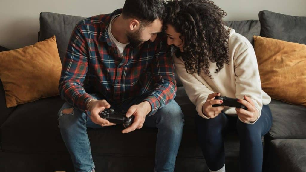 A couple sitting on a couch smiling while playing video games with controllers.