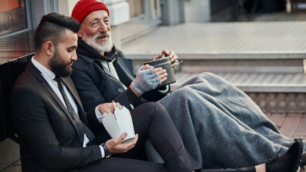 A bearded man in a black suit is having a snack with an old person while sitting on a bench.