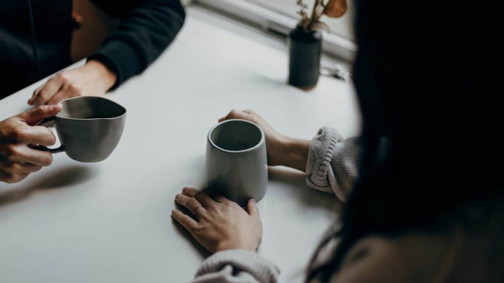 Two people's hands are visible around gray ceramic mugs on a white table, suggesting a conversation over coffee or tea.