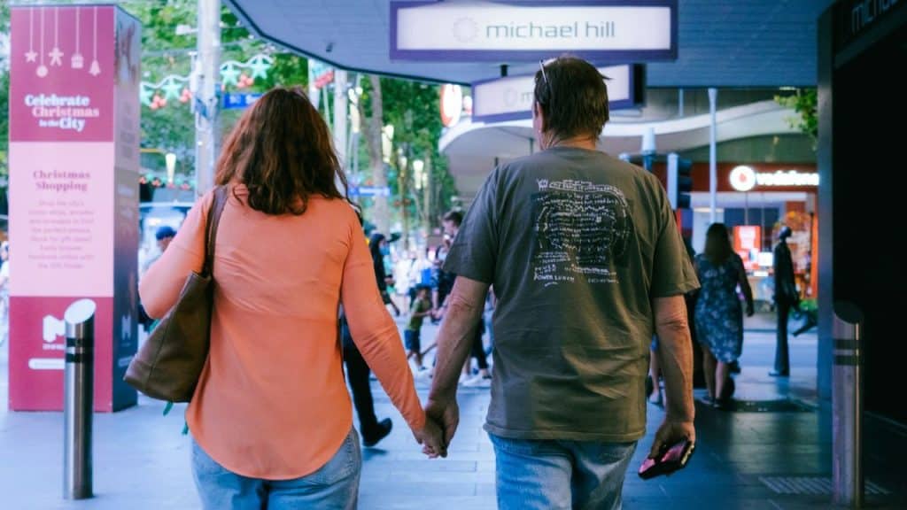 A man and woman, seen from behind, hold hands while walking through a city shopping area.