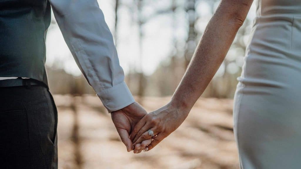 A couple holding hands outdoors, one wearing a wedding ring.