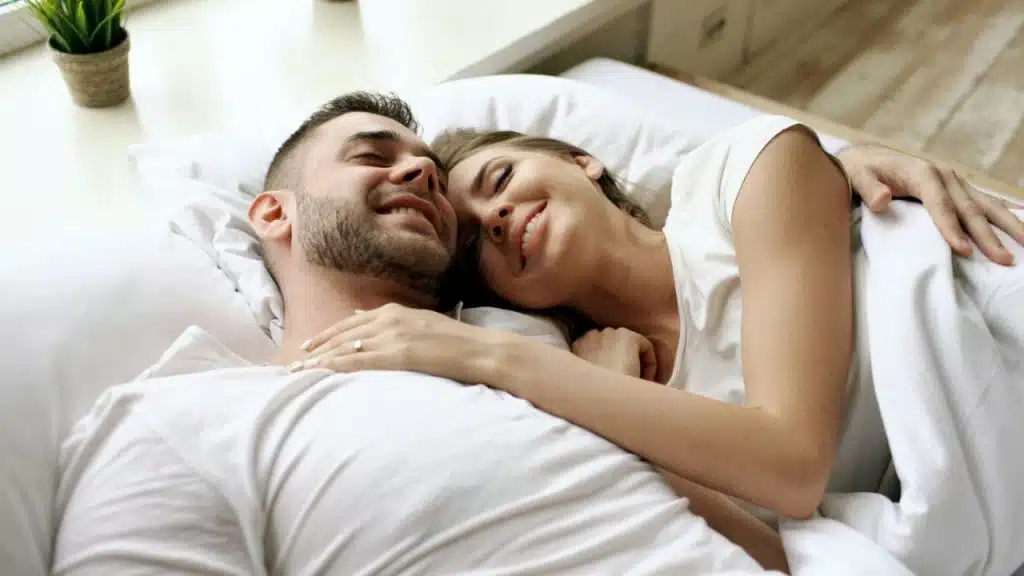 A smiling man and woman are lying together in a bright bed with white bedding.