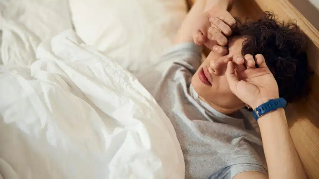 A person with short curly hair is lying in bed, rubbing their eyes.