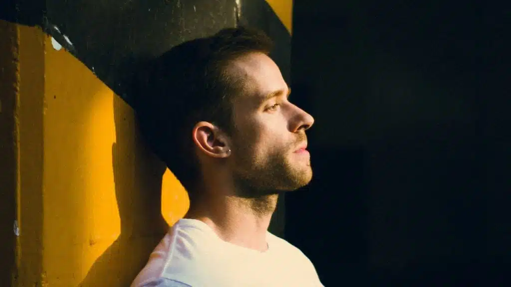 A man in a white shirt is leaning against a yellow and black wall, looking up to the side.