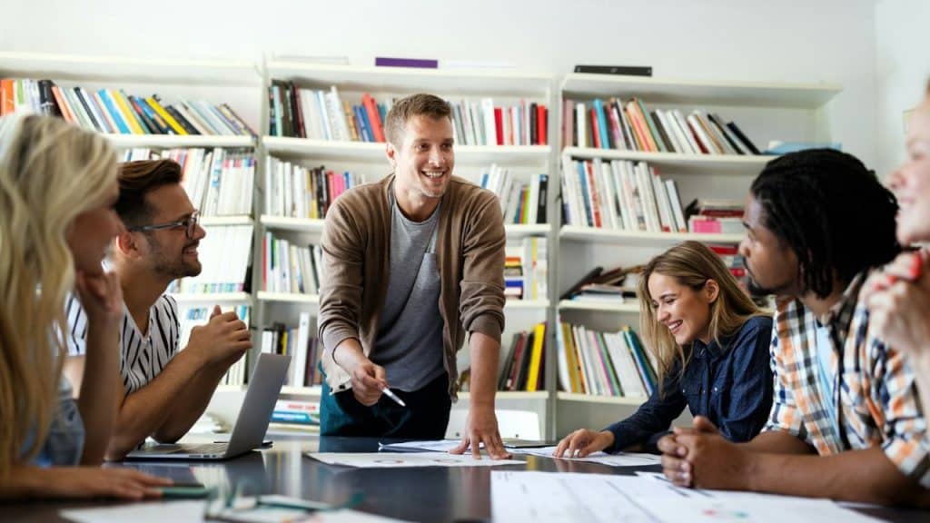 Six young adults gathered around a table with paperwork in a room with a bookshelf.