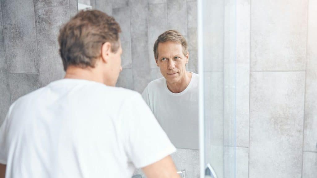Middle-aged man in a white shirt looking at his reflection in a bathroom mirror.