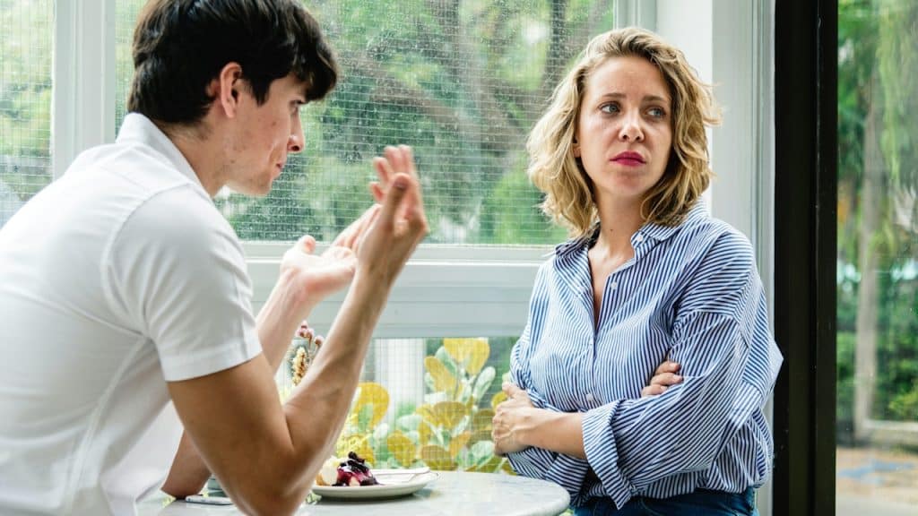A blonde woman with her arms crossed listens with a serious expression as a man gestures while talking to her at a table.