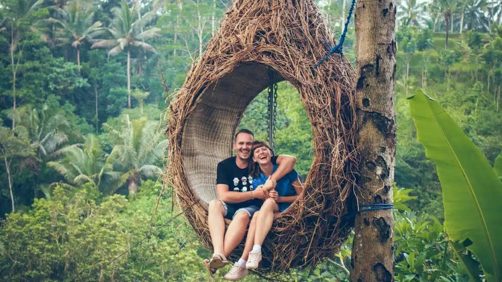 A man and woman riding on a brown hammock beside a brown tree.