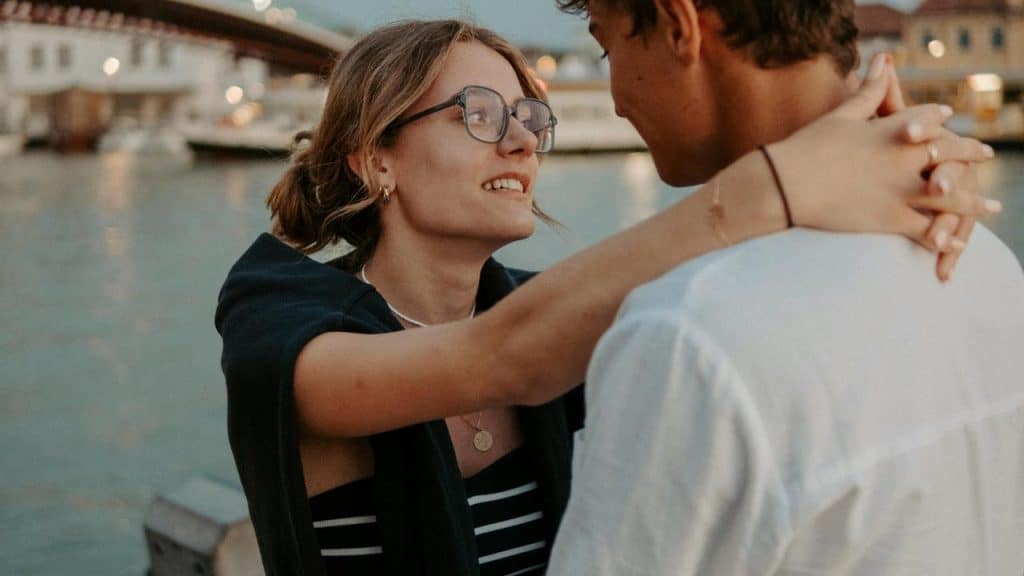 A woman smiling and embracing a man near the water at sunset.