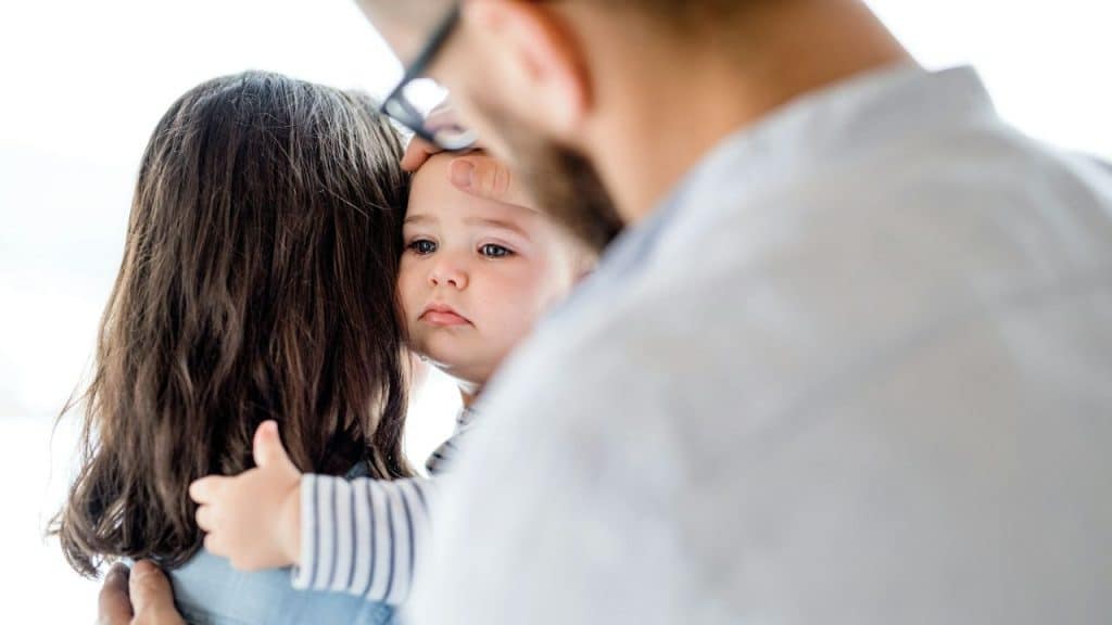 A child is being held by a woman while a man gently touches their forehead.