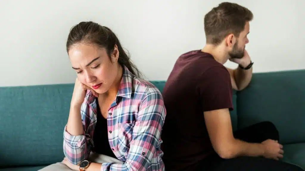 A couple sits back-to-back on a teal couch, looking away from each other in silence.