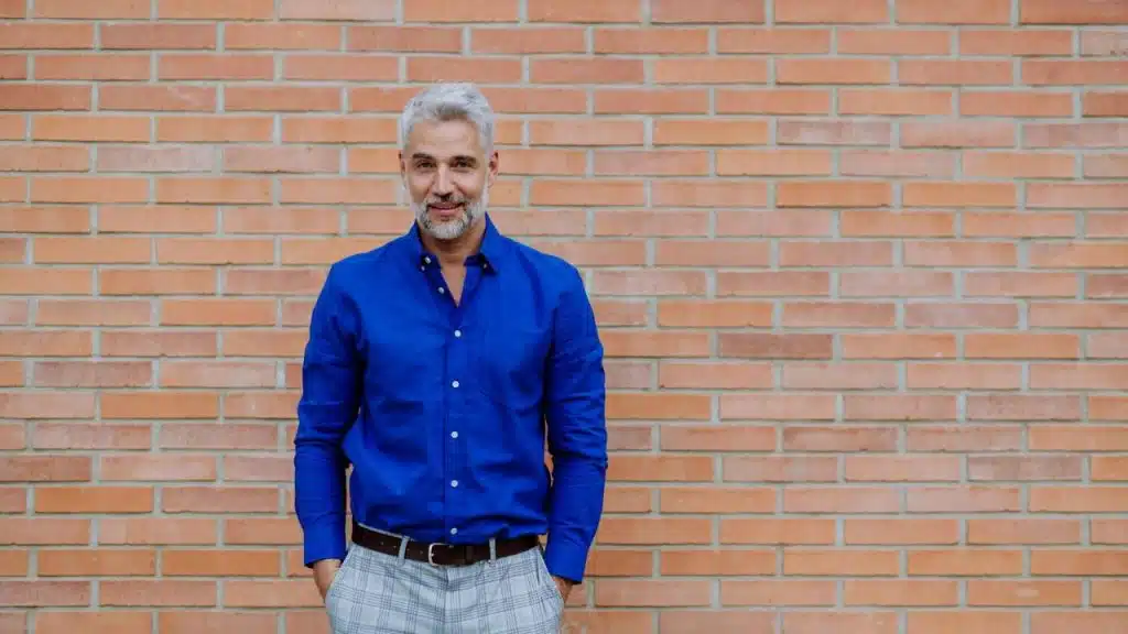 A gray-haired man in a blue shirt and plaid pants stands against a brick wall.