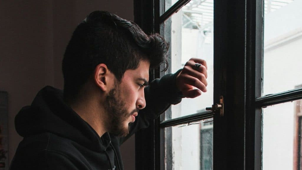 A bearded man in a black hoodie is looking out a dark-framed window.