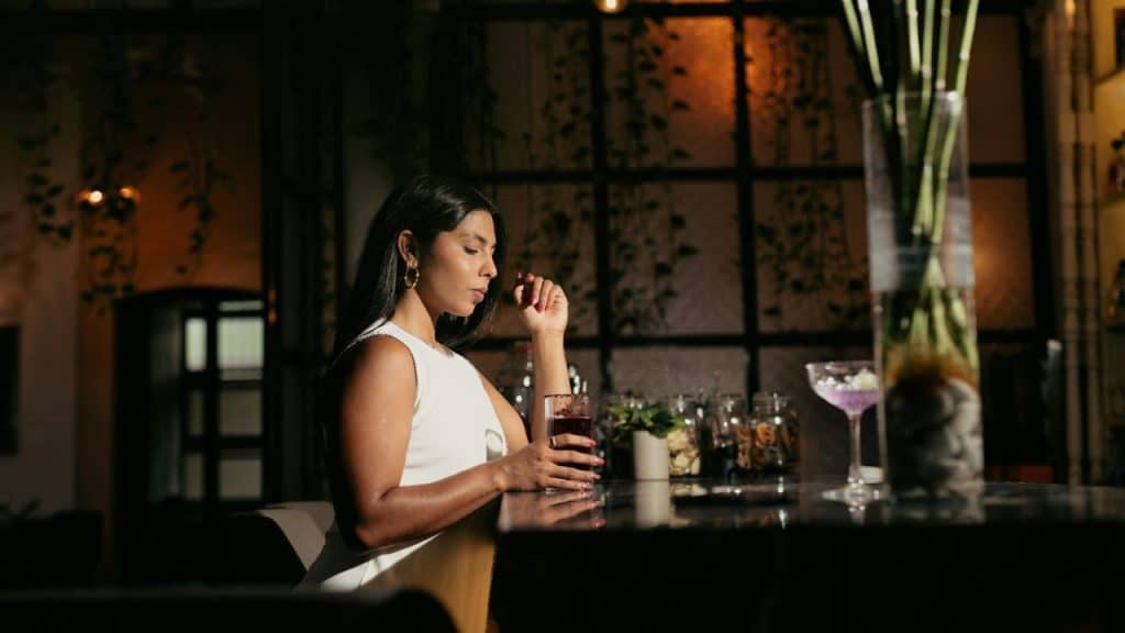 A woman in a white dress is sitting alone at a bar, holding a dark red drink.