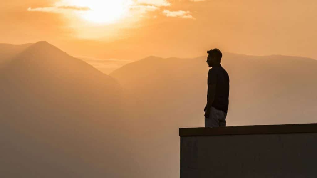 A silhouetted man stands on a rooftop looking at hazy, sunlit mountains.