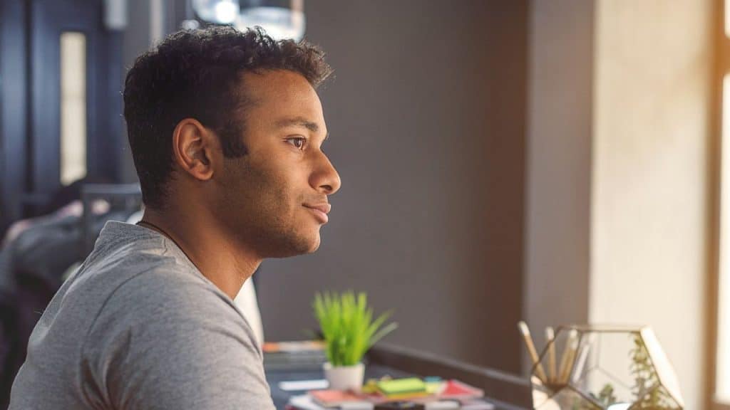 Man in a gray shirt looking to the side in a sunlit room, with a small potted plant on the desk in front of him.