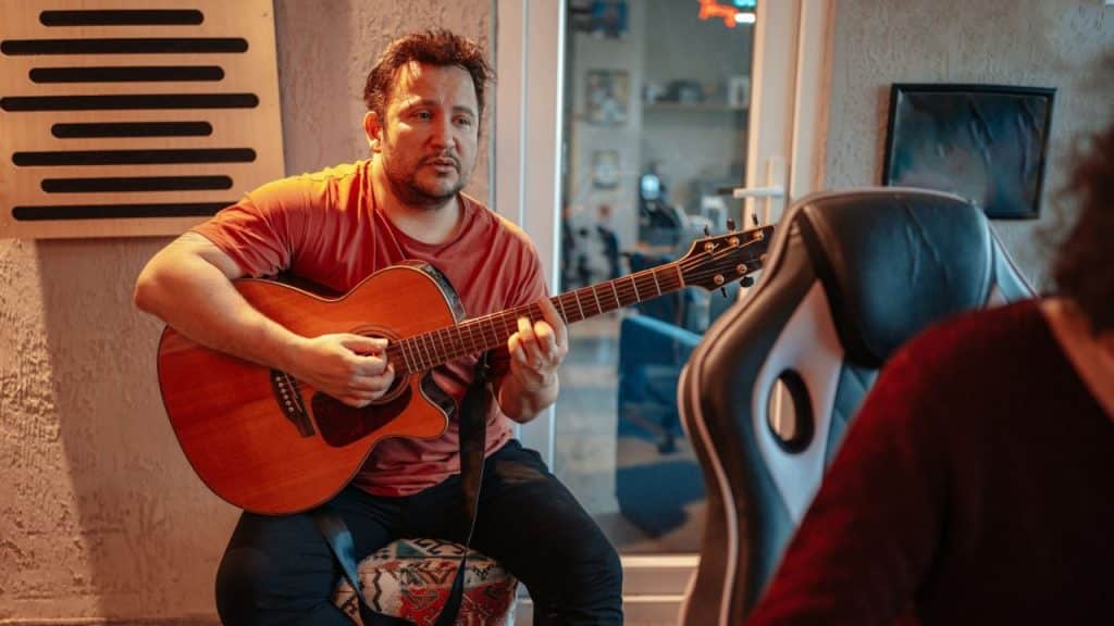 Man in an orange shirt sitting and playing an acoustic guitar indoors, facing a person whose back is visible.
