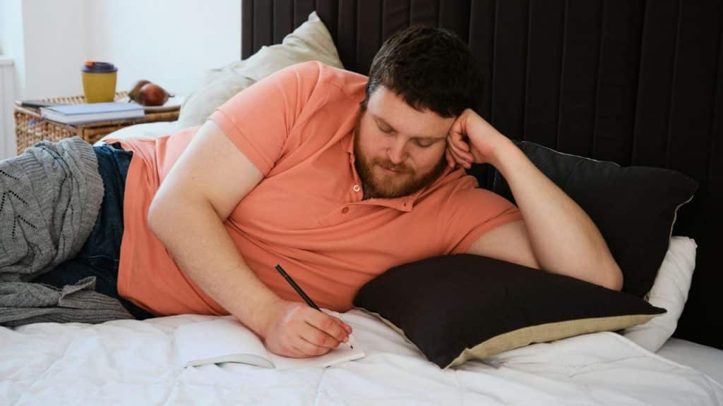 A bearded man is lying on a bed and writing in a notebook with a pencil.