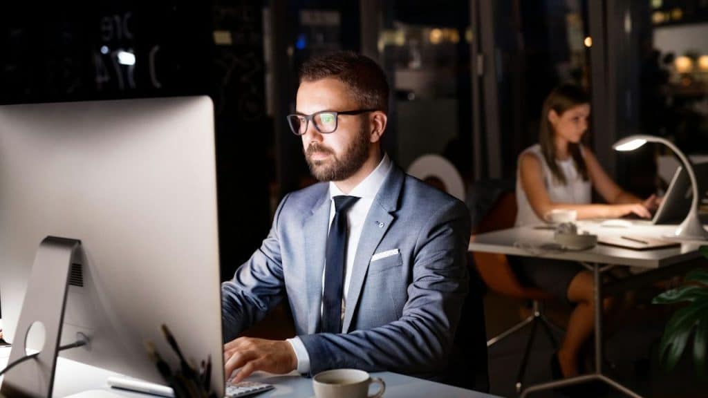 Man in suit and glasses works on a large computer monitor in a dark office.