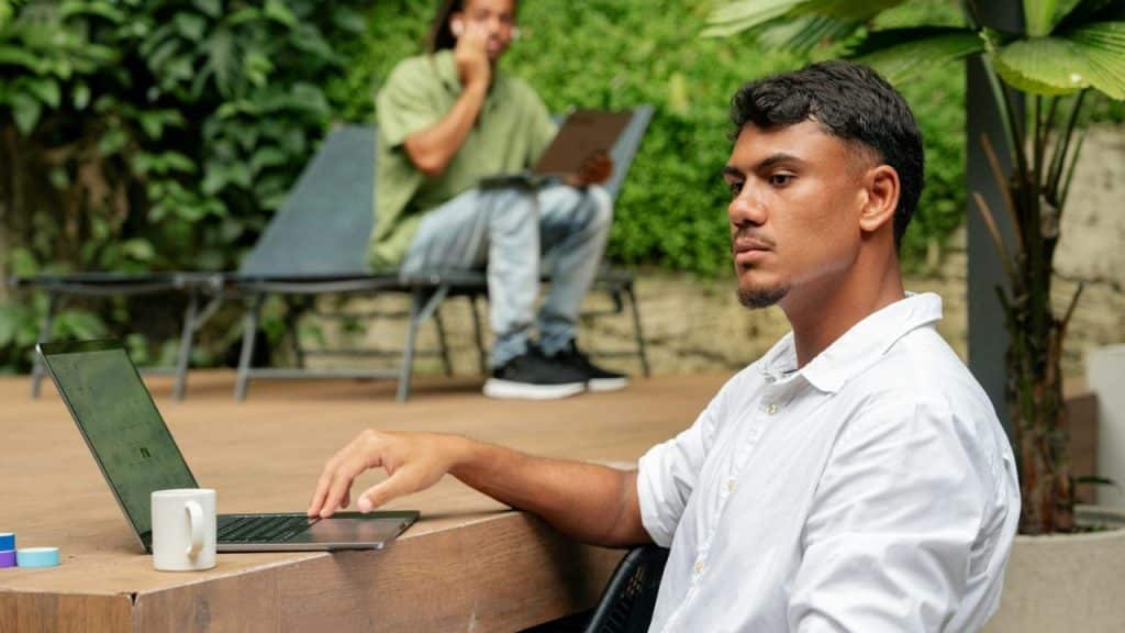 Young man in white shirt sits at a table with a laptop, outdoors, with another man in the background.