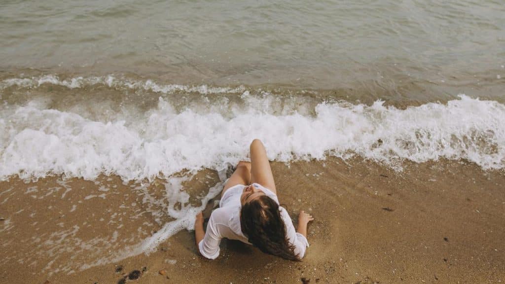 A woman sitting on the shore with waves gently washing over her legs.