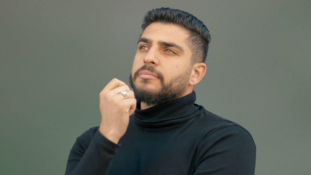 Man in a black turtleneck and beard, holding his chin, looking up with a thoughtful expression.