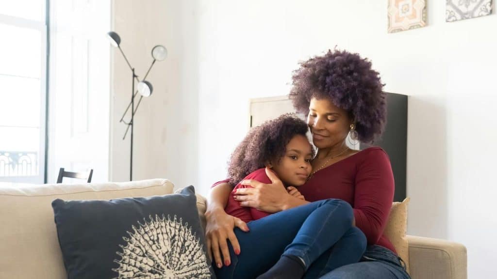 A mother sitting on a couch hugging her daughter.