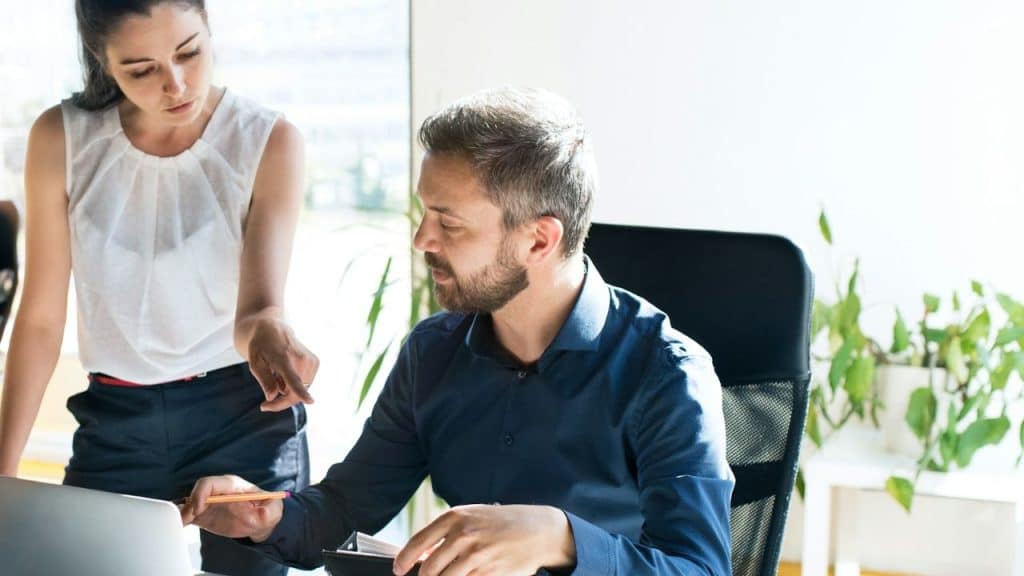 A man and a woman are standing next to a desk, working together in a sunny office.