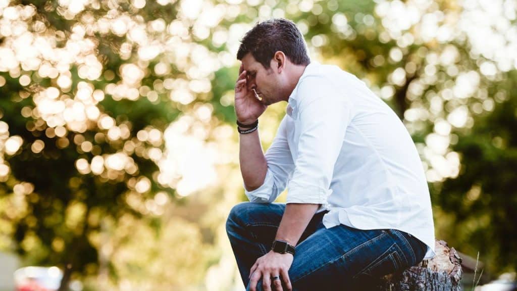 A pensive man in a white shirt and jeans sits on a log, holding his forehead.