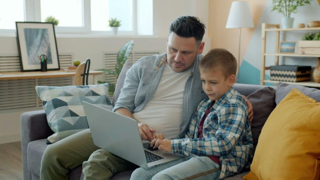 Man and boy sitting on a couch, looking at and using a laptop together.