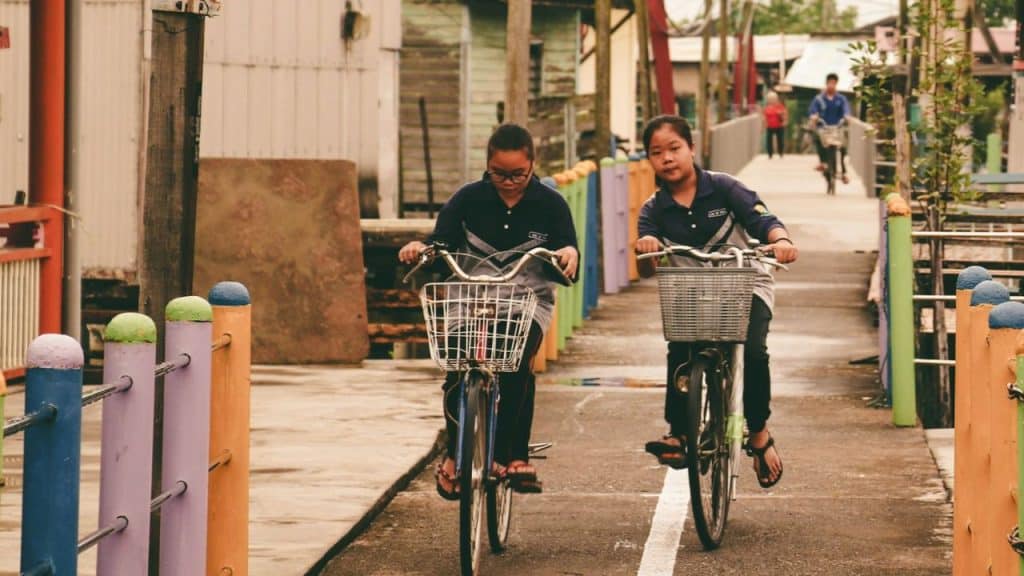 Two children riding bicycles side-by-side down a colorful wooden walkway outdoors.
