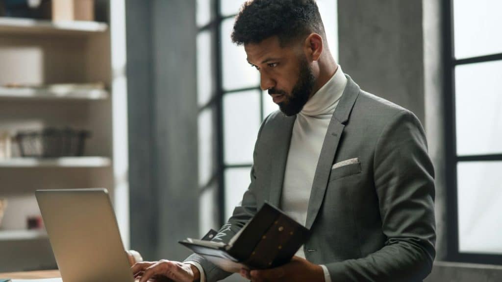 Man in a turtleneck and gray suit jacket working on a laptop while holding a notebook.