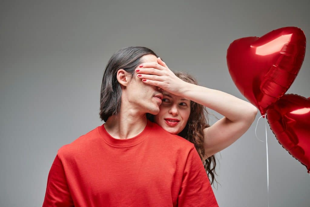 A woman covering the eyes of the man holding heart-shaped balloons