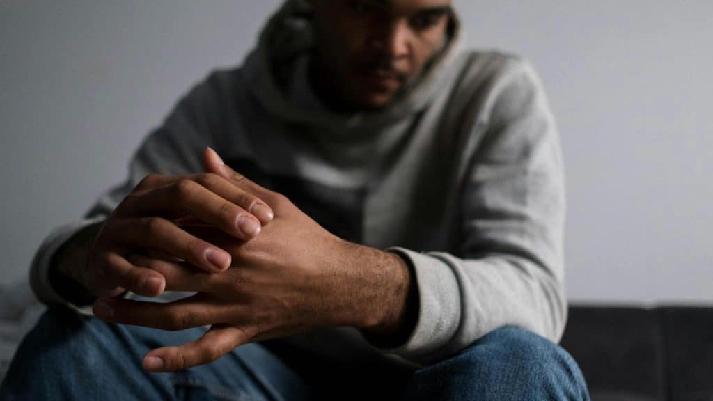 A man in a hoodie sits with clasped hands, looking down thoughtfully.