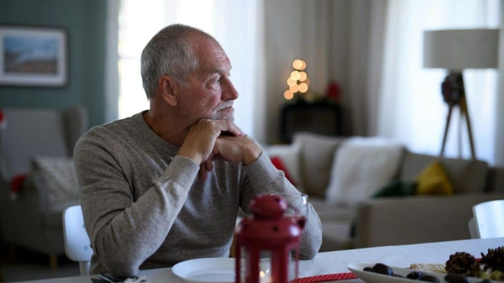 Older man sitting alone at a table with his chin resting on his hands.