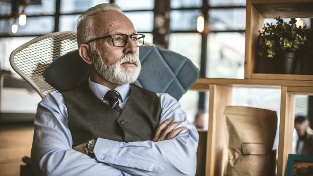 Older man with a white beard, glasses, and a vest sitting with arms crossed.