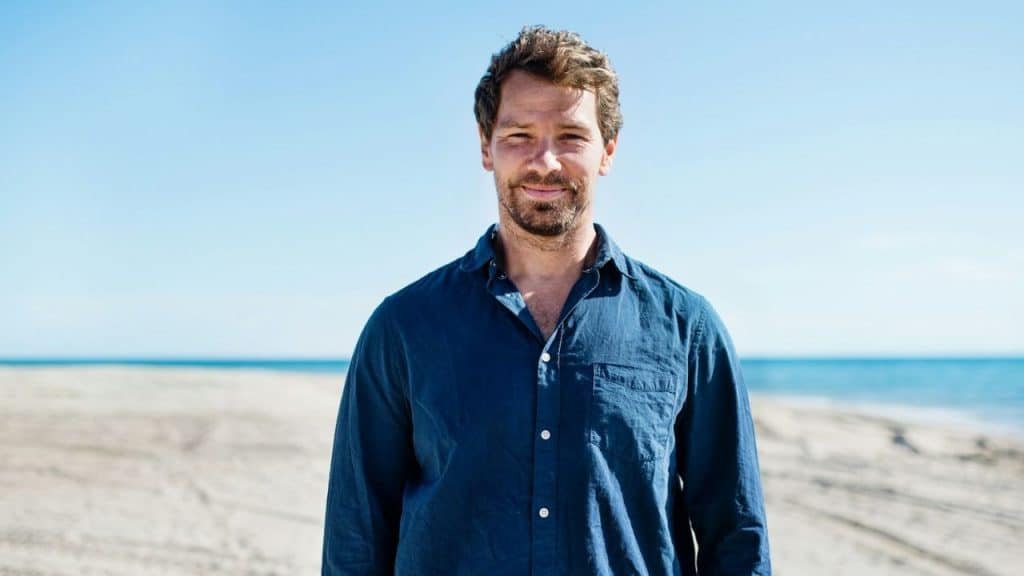 Close-up of a smiling man in a blue shirt standing on a sandy beach.