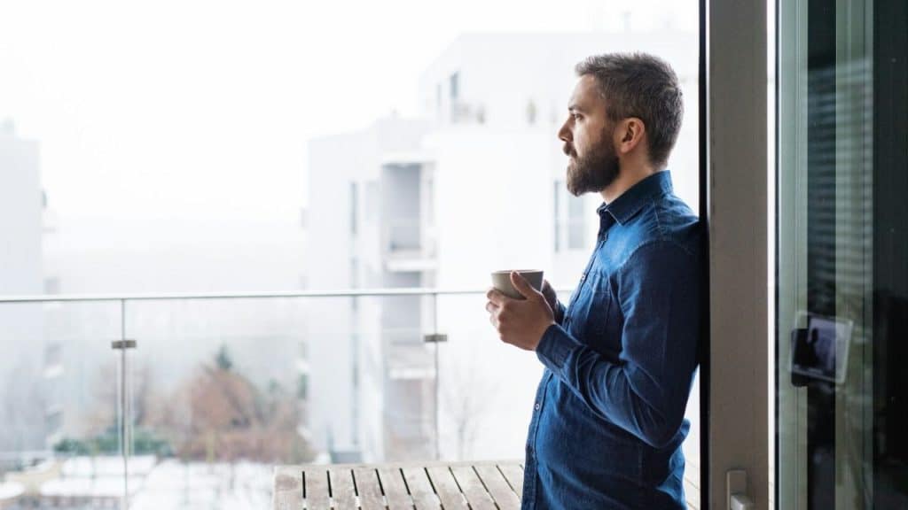 Man with a beard holding a cup, leaning on a balcony door in an urban setting.