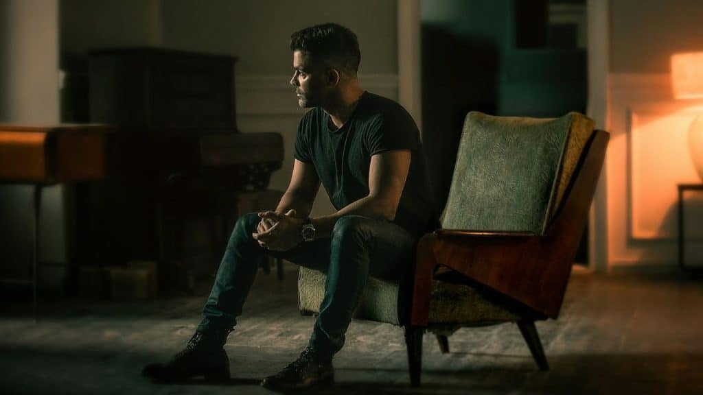 Man in a dark t-shirt and jeans sitting alone in a mid-century armchair.