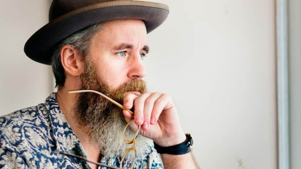 Man with a full beard, hat, and patterned shirt looking thoughtfully to the side.