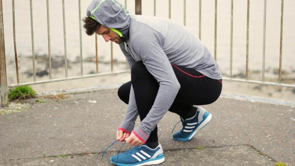 A man in athletic wear squatting down to tie the laces of a blue running shoe.