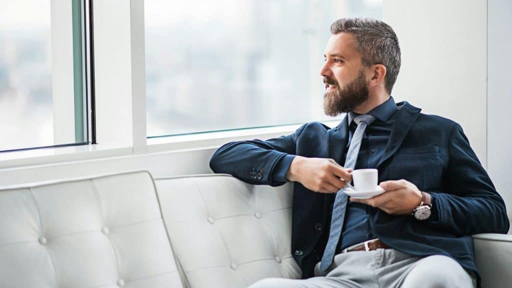 Bearded man in a suit and tie sitting on a white couch, holding a cup.