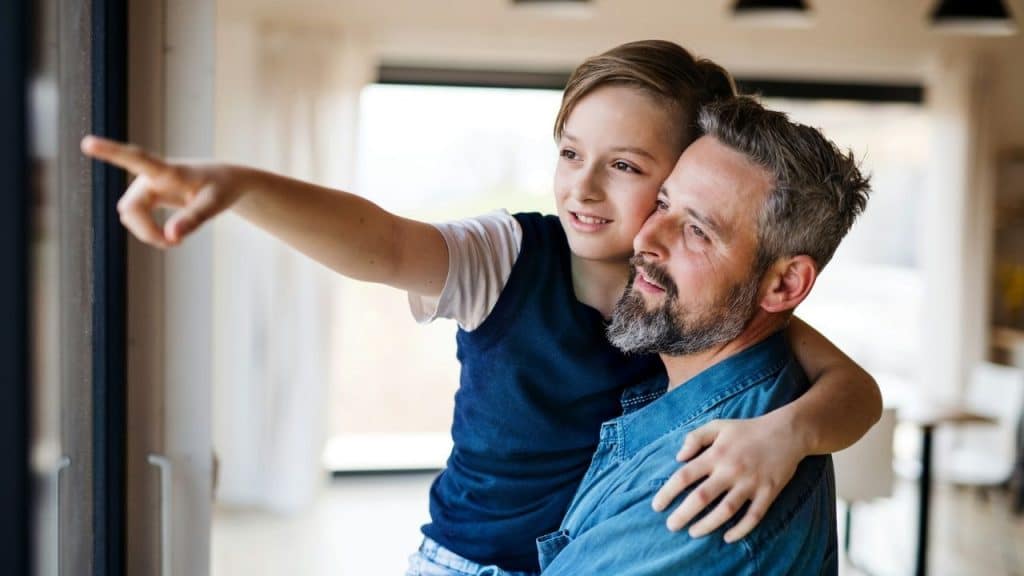 Father and son embracing indoors, the son pointing out a window.