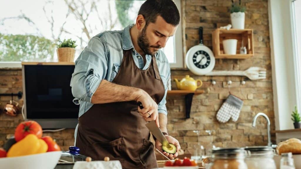 A man in an apron is cutting an avocado in a bright, rustic kitchen.