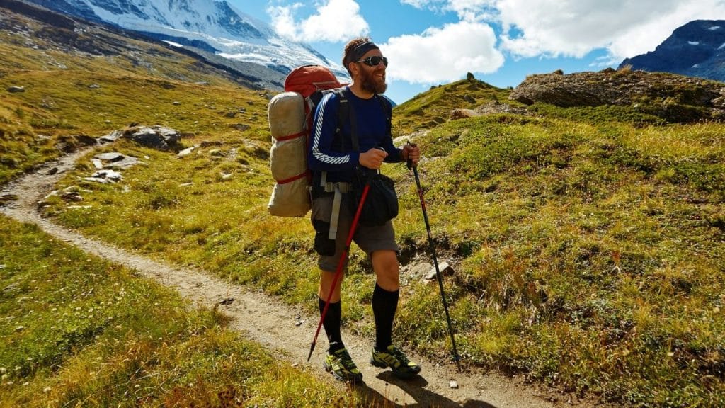 Bearded hiker with large backpack and trekking poles on a mountain trail.