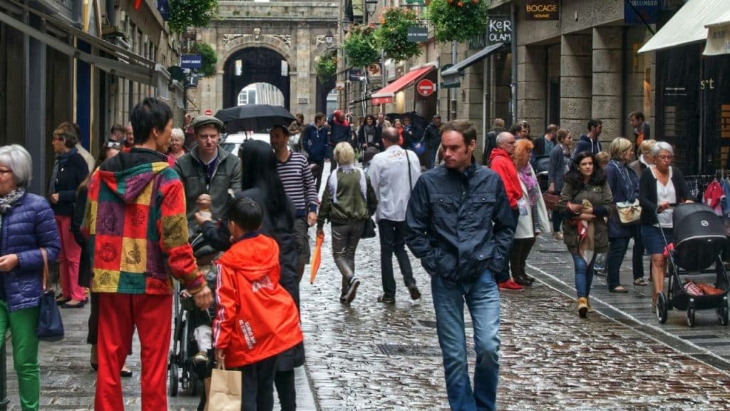Crowded, rainy cobblestone street with pedestrians, shops, and an arched entryway.