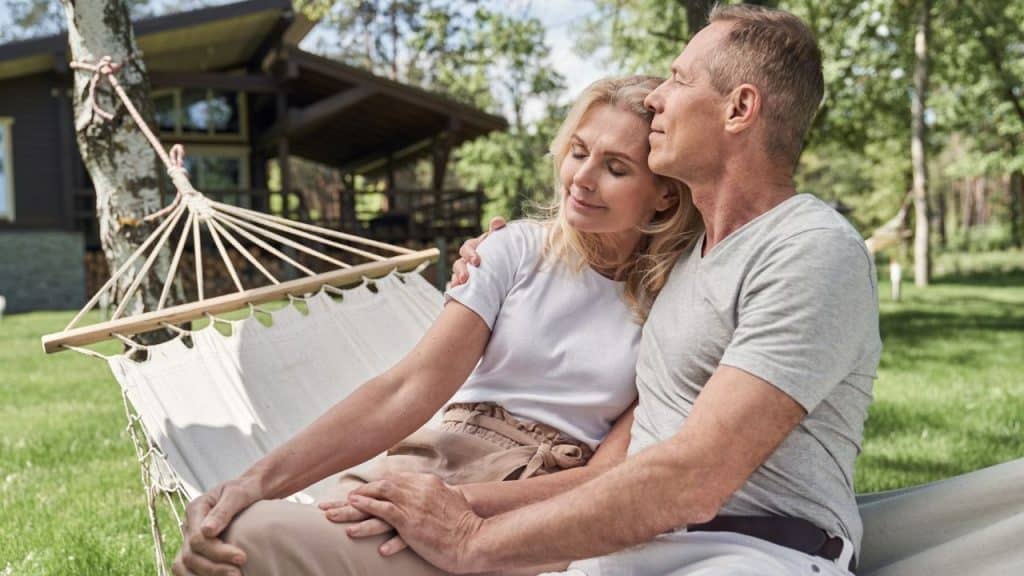 Middle-aged couple embracing and relaxing in a hammock outside a wooden house.
