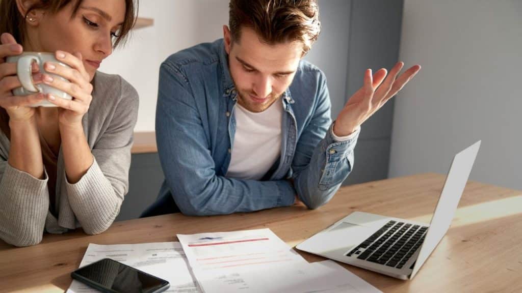 Concerned couple reviewing bills and documents at a table with a laptop and a mug.