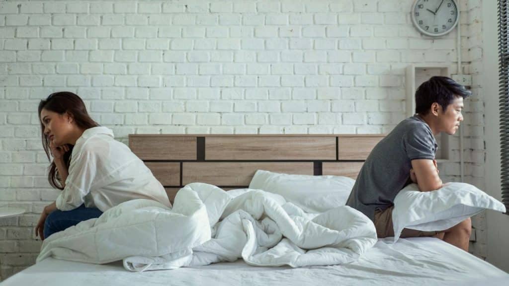 Unhappy couple sitting with their backs to each other on a bed with white brick wall.