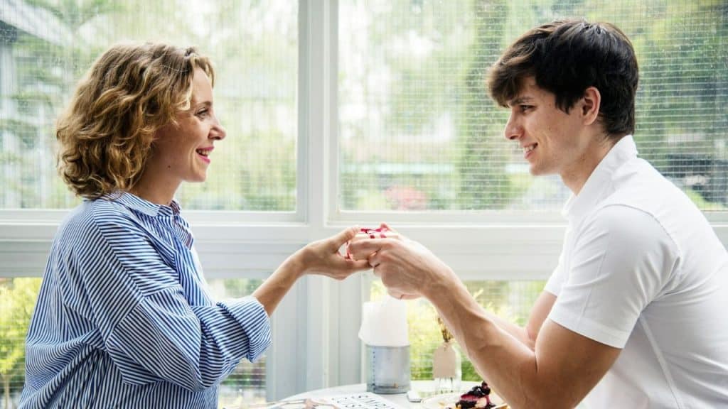 Smiling couple sitting at a table near a window, holding hands over a small box.